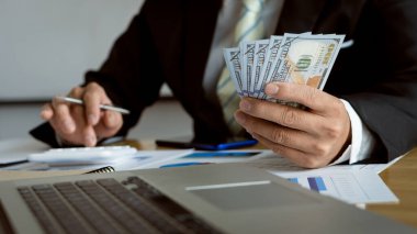 Asyalı işadamı, hesap makinesi ve dizüstü bilgisayarla para sayma kavramı ile ahşap masada banknotları sayıyor. Maaş. Şirket ofisinde çalışanlara ödeme yapmak.