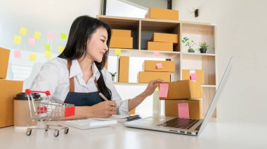 Küçük işletme girişimi, KME sahibi, Asyalı bayan girişimci okuyun ve sipariş numarasını dizüstü bilgisayara yazın. Müşterilere satılan kutuları hazırlamak için çevrimiçi siparişleri kontrol et. Çevrimiçi KOBİ iş fikri.