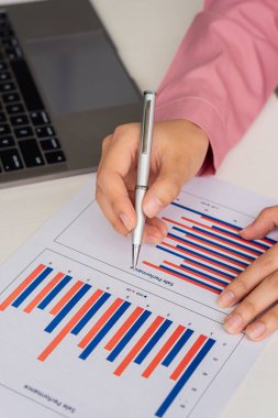 Genç bir finans analisti, ahşap bir masada otururken dizüstü bilgisayarında çalışıyor. Eldeki belgeleri analiz eden işadamı dikey resimli grafik ve finansal konsept hesap makinesi.