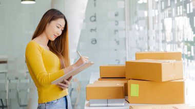 Güzel bir Asyalı iş kadını müşterilerden sipariş almak için kutular ve dizüstü bilgisayarlarla çalışır ve evde çalışmaktan zevk alır. Fikir satar ve dağıtırlar..