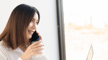 Genç bir kadın dizüstü bilgisayarın önünde çalışırken telefonla konuşuyor ve kahve içiyor.