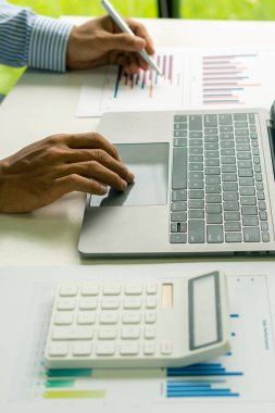 Genç bir finans analisti ahşap bir masada otururken ofiste çalışır. İş adamı elindeki grafik belgesini analiz ediyor ve bir sayı makinesi ve dizüstü bilgisayarla hesaplıyor