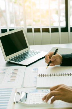 Genç bir finans analisti ahşap bir masada otururken ofiste çalışır. İş adamı elindeki grafik belgesini analiz ediyor ve bir sayı makinesi ve dizüstü bilgisayarla hesaplıyor