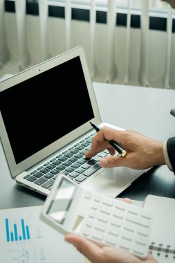 Genç bir finans analisti ahşap bir masada otururken ofiste çalışır. İş adamı elindeki grafik belgesini analiz ediyor ve bir sayı makinesi ve dizüstü bilgisayarla hesaplıyor