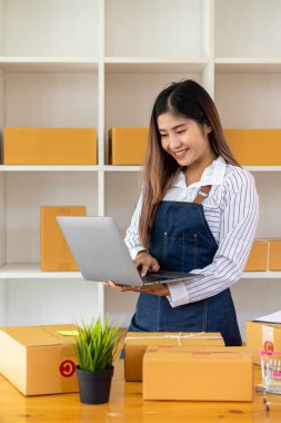 Küçük bir işletme kuran KOBİ girişimcisi, evde serbest meslek sahibi Asyalı bir kadının portresi, Kutular, Smartphone, Laptop, Çevrimiçi Pazarlama, Paketleme, KME Nakliyat, E-ticaret ve Dağıtım Fikirleri.