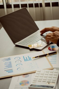 Genç işadamı masadaki iş grafiği belgelerini dizüstü bilgisayar ve hesap makinesi ile analiz ediyor. İş dünyası ve dikey resimli ekonomik kavramlar.