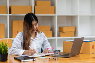 Evde internet üzerinden çalışan genç iş kadını. Kadın işadamları evde çalışmak ve KOBİ işine başlamak için dizüstü bilgisayar kullanıyorlar. İş yerinde küçük ve karton kutular var.