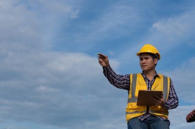 Asyalı erkek endüstriyel mühendis sarı bir şapka takıyor ve Pam kağıt tutarak merdivenlerde oturuyor ve gökyüzü endüstriyel mühendislik kavramının arkasına işaret ediyor.
