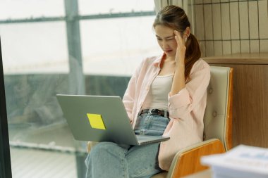 Asyalı endişeli bir kadın dizüstü bilgisayarı ile cam kenarında oturur, muhtemelen dizüstü bilgisayarla sürekli çalışmaktan kaynaklanan stresten..