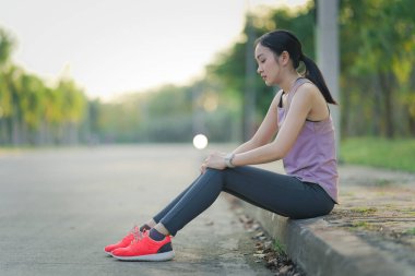 Bir kadın çimlerin üzerinde otururken bileğini acı içinde tutuyor. Buna yaralanma, burkulma, ayak bileği ağrısı ya da dışarıda egzersiz yaparken meydana gelen bir kaza neden olabilir..