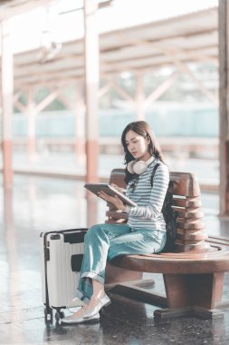 Güzel Asyalı bir kadın, elinde defteriyle tren istasyonundaki ahşap bankta dinleniyor. Sırt çantasıyla tren bekliyor. Seyahat yaşam tarzının tadını çıkarıyor..