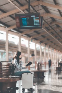 Güzel Asyalı bir kadın, elinde defteriyle tren istasyonundaki ahşap bankta dinleniyor. Sırt çantasıyla tren bekliyor. Seyahat yaşam tarzının tadını çıkarıyor..