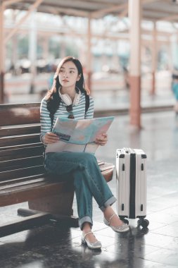 Güzel Asyalı bir kadın, elinde defteriyle tren istasyonundaki ahşap bankta dinleniyor. Sırt çantasıyla tren bekliyor. Seyahat yaşam tarzının tadını çıkarıyor..