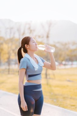 Asyalı bir kadın parkta egzersiz yaptıktan sonra su içer..