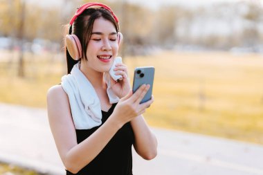Asyalı bir kadın kulaklık takıyor, alnındaki teri siliyor ve egzersiz yaptıktan sonra telefonuna bakıyor..
