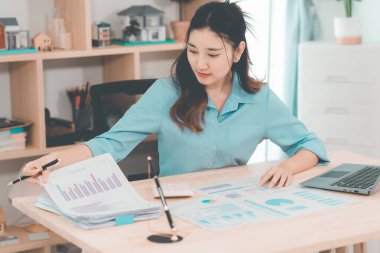 Asyalı bir kadın muhasebeci masasında oturup dizüstü bilgisayarında dosya ve mali belgeler okuyor, strateji ve planlamaya odaklanıyor..
