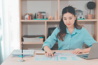 Asyalı bir kadın muhasebeci masasında oturup dizüstü bilgisayarında dosya ve mali belgeler okuyor, strateji ve planlamaya odaklanıyor..