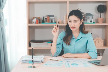 Asyalı bir kadın muhasebeci masasında oturup dizüstü bilgisayarında dosya ve mali belgeler okuyor, strateji ve planlamaya odaklanıyor..