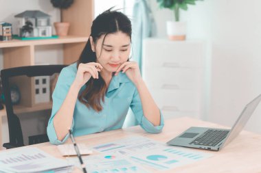 Asyalı bir kadın muhasebeci masasında oturup dizüstü bilgisayarında dosya ve mali belgeler okuyor, strateji ve planlamaya odaklanıyor..