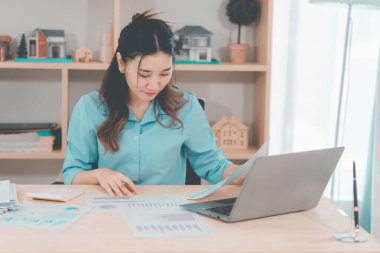 Asyalı bir kadın muhasebeci masasında oturup dizüstü bilgisayarında dosya ve mali belgeler okuyor, strateji ve planlamaya odaklanıyor..