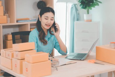 Asian woman ships packages using a laptop. E-commerce order management, logistics arrangements, shipping concept, SME business.