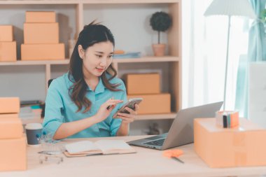 Asyalı bir kadın dizüstü bilgisayar kullanarak paket taşıyor. E-ticaret sipariş yönetimi, lojistik düzenlemeler, nakliye konsepti, KOBİ işi.