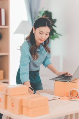 Asian woman ships packages using a laptop. E-commerce order management, logistics arrangements, shipping concept, SME business.