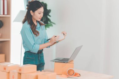 Asian woman ships packages using a laptop. E-commerce order management, logistics arrangements, shipping concept, SME business.