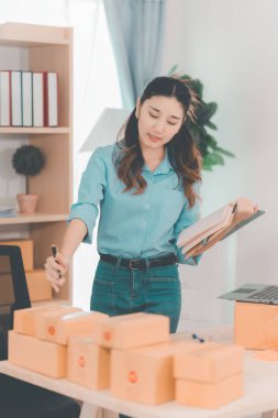 Asian woman ships packages using a laptop. E-commerce order management, logistics arrangements, shipping concept, SME business.