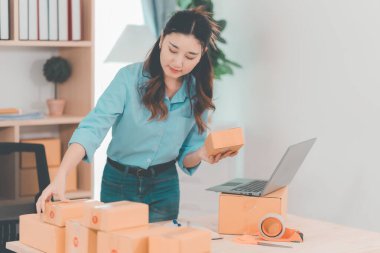 Asian woman ships packages using a laptop. E-commerce order management, logistics arrangements, shipping concept, SME business.