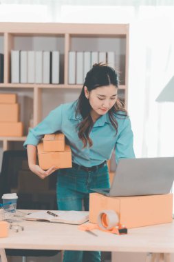 Asian woman ships packages using a laptop. E-commerce order management, logistics arrangements, shipping concept, SME business.