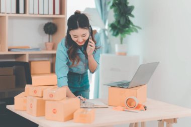 Asyalı bir kadın dizüstü bilgisayar kullanarak paket taşıyor. E-ticaret sipariş yönetimi, lojistik düzenlemeler, nakliye konsepti, KOBİ işi.