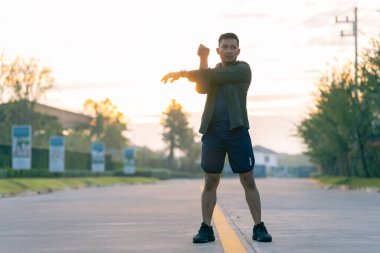 Tişörtlü ve şortlu genç bir adam geriniyor. Arka planda güneş ışığı parlıyor. Antrenman öncesi atmosferi yaratıyor..