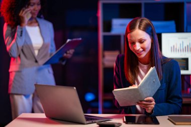 İki neşeli iş kadını, gece geç saatlere kadar çalışırken iş raporlarını meslektaşlarına açıklamak için yoğun bir şekilde veri analizi ve fikir alışverişinde bulunuyorlar..