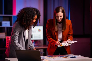 İki neşeli iş kadını, gece geç saatlere kadar çalışırken iş raporlarını meslektaşlarına açıklamak için yoğun bir şekilde veri analizi ve fikir alışverişinde bulunuyorlar..