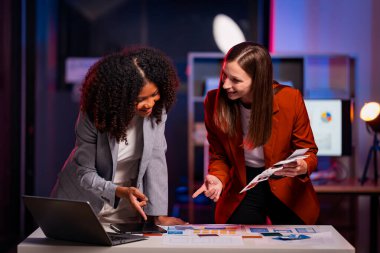 İki neşeli iş kadını, gece geç saatlere kadar çalışırken iş raporlarını meslektaşlarına açıklamak için yoğun bir şekilde veri analizi ve fikir alışverişinde bulunuyorlar..
