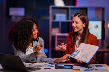 İki neşeli iş kadını, gece geç saatlere kadar çalışırken iş raporlarını meslektaşlarına açıklamak için yoğun bir şekilde veri analizi ve fikir alışverişinde bulunuyorlar..