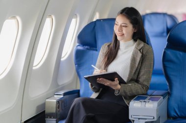 An Asian woman is using a digital tablet while traveling on an airplane near the window. She is traveling for business.