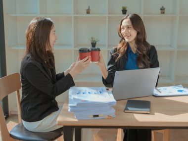 İki Asyalı iş kadını taşınabilir kahve fincanlarını tutuyor ve iş başarılarını kutlamak için onları birbirine tokuşturuyorlar; kağıt kahve fincanlarını tokuşturma işi..