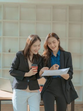 İki Asyalı iş kadını birlikte çalışırken resmi verileri tartışıyor, finansal tablolar ve bilgileri bir dizüstü bilgisayar ekranında ve modern ofis belgelerinde tartışıyorlar.