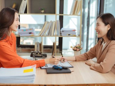 İki Asyalı iş kadını gülümsüyor ve sohbet ediyor, ellerinde iş anlaşmaları veya araba satışları veya sigorta danışmanlığı için belgeleri imzalamaya hazır kalemler tutuyorlar..