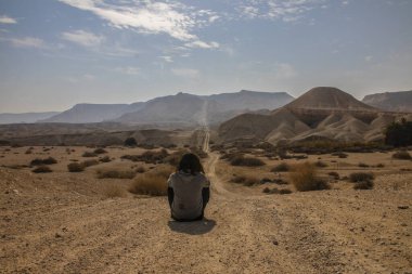 Genç adam çölün ortasında oturuyor ve uzun bir yola bakıyor.