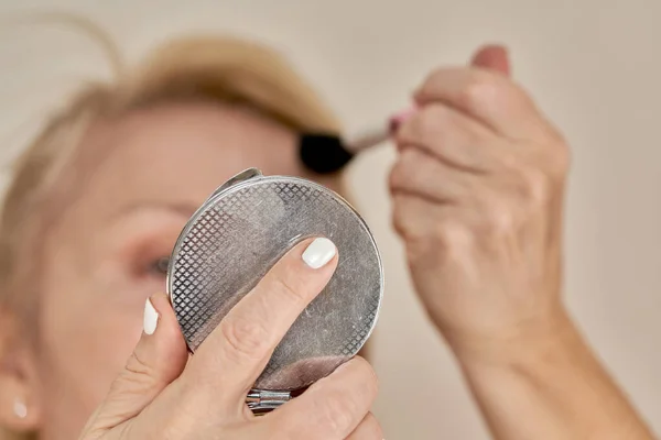 A woman with a cosmetic brush applies makeup to her face while looking in the mirror. Cosmetic skin care