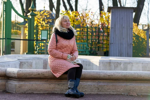 一个金发碧眼 身穿粉色外套的女人坐在一个无法使用的喷泉石墙上 — 图库照片