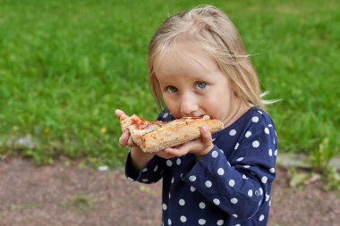 Beyaz puantiyeli mavi elbiseli küçük kız yeşil çimenlerin üzerinde büyük bir dilim pizza yiyor.