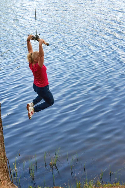 スポーツウェアを着た中年女性が水の上にロープで棒にぶら下がっています 自然の中でスポーツ — ストック写真