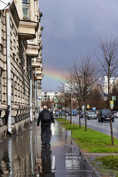 男は雨の後にぬれた通りを歩いて空に虹があり 後ろから見る — ストック写真