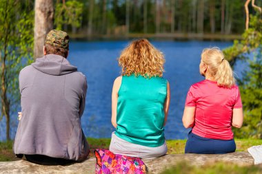 Bir adam ve iki kadın sırtı kameraya dönük bir şekilde bir orman gölünün kıyısında oturuyorlar. İnsanlar göle hayran.