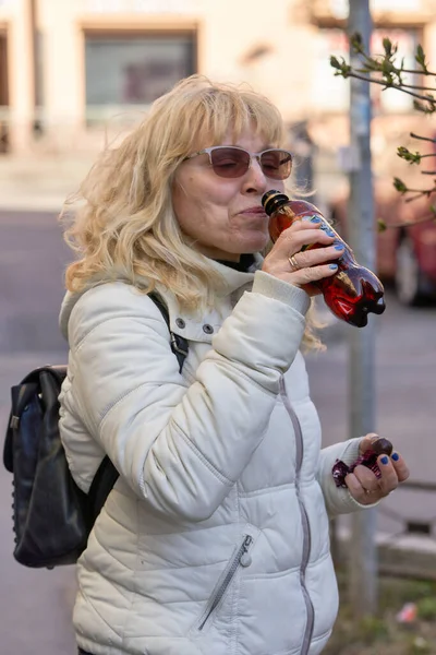一位戴太阳镜的金发老妇人走在街上 喝着瓶子里的水 — 图库照片