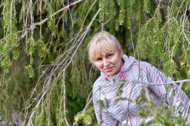 Bir köknar ağacının dalları arasında sarı saçlı orta yaşlı bir kadının portresi.